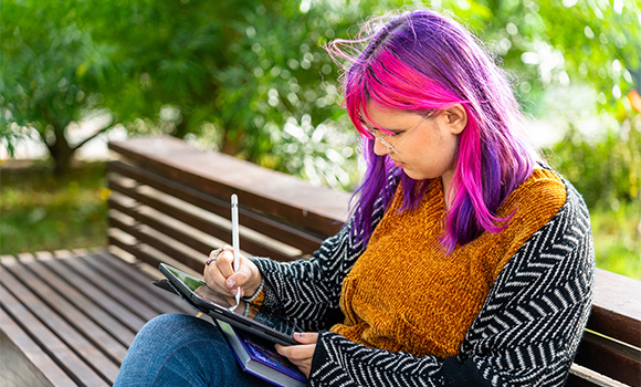 young person with purple and pink hair sitting on a bench using a tablet to draw or write in a park showcasing creativity in 2023