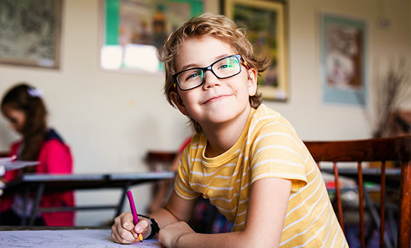 smiling boy wearing glasses in classroom working on schoolwork with colorful surroundings and classmates focusing on learning 22 educational activities