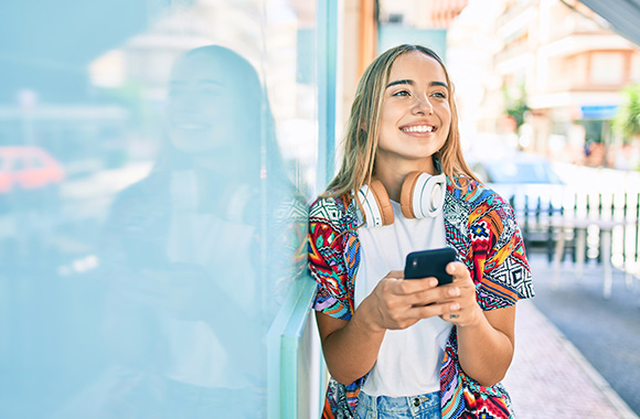 young woman with headphones smiling while using smartphone on a city street featuring vibrant colors showing a sense of joy and youthful energy associated with age 13 and digital connectivity