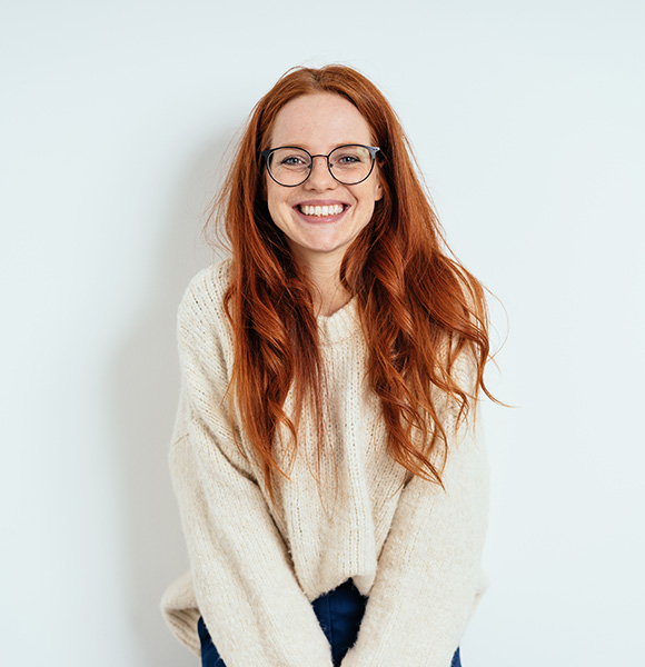 smiling woman with long red hair wearing glasses and a cozy sweater standing against a light background reflecting joy and positivity associated with nine attributes